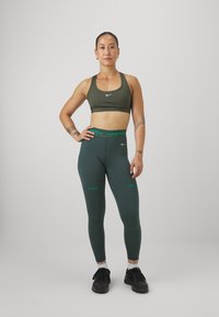 Tmavě zelená sportovní podprsenka a odpovídající legíny s zeleným logem "NIKE PRO". Modelka má na sobě černé tenisky a bílé ponožky.
