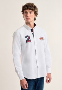 Camisa blanca de manga larga con cuello abotonado, que presenta un "2" a cuadros y bordado de caballo en colores contrastantes. Textura de algodón suave.