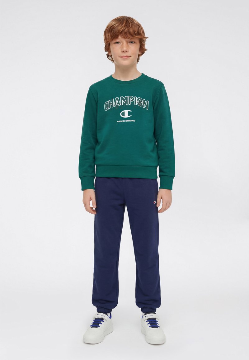 Jongen met rood haar die een groen Champion-sweatshirt, marineblauwe joggingbroek en witte sneakers draagt, staand tegen een effen witte achtergrond.