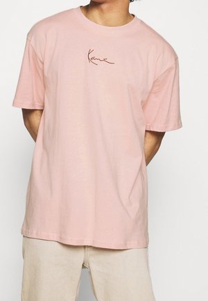 Person trägt ein locker sitzendes hellrosa kurzärmeliges T-Shirt mit kleinem braunen Schriftzug in der Mitte der Brust und hellbeige Hose.
