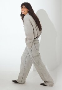 Camisa de manga larga oversize en gris claro combinada con pantalones vaqueros de pierna ancha en gris claro. Los zapatos tienen un diseño de punta afilada en negro.