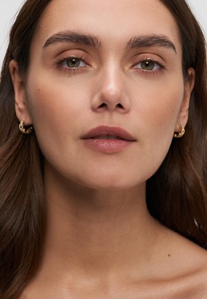 Junge Frau mit langen braunen Haaren, die kleine goldene Creolen trägt, blickt mit neutralem Ausdruck direkt nach vorne vor einem einfarbigen Hintergrund.