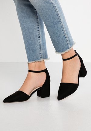 Voeten met zwarte gesloten schoenen met blokhak en enkelriempjes, gecombineerd met lichtblauwe spijkerbroeken met rafelige zoom.