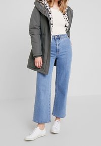 Oliwkowa ocieplana kurtka z podszewką z futerka w panterkę, zestawiona z wysokim stanem szerokimi jasnoniebieskimi dżinsami i białymi trampkami.