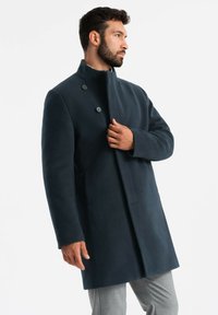 Cappotto lungo in lana blu scuro con colletto alto, chiusura a doppio bottone e una silhouette dritta. Texture liscia, senza motivi visibili.