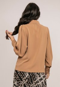 Blouse beige avec de longues manches bouffantes, un col classique et des poignets boutonnés. Portez-la sur une jupe à motifs noirs et beiges. Texture lisse.