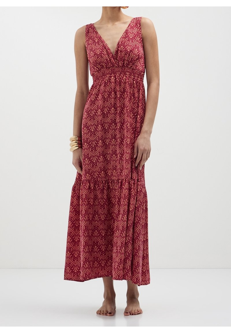 Femme porte une robe maxi rouge sans manches avec un motif abstrait rose clair et un décolleté en V profond, debout pieds nus sur un fond uni.