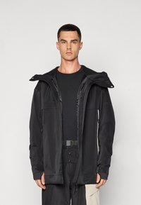Casaco impermeável preto com capuz, bolsos com zíper e um design elegante. Tecido leve com uma textura suave.