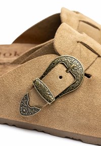 Sandalias tipo slip-on de ante marrón claro con una hebilla decorativa de bronce con patrones florales y detalles texturizados.