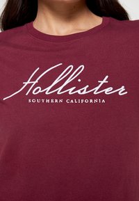 Camiseta de manga corta color burdeos, hecha de algodón, con texto en cursiva blanca que dice "Hollister" y "SOUTHERN CALIFORNIA" debajo.