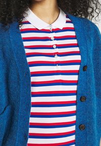 Modrý chlupatý cardigan přes bílou a červenou pruhovanou polo košili. Polo košile má límec a knoflíky; cardigan má velké tmavé knoflíky. Měkká textura.