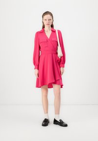 Abito avvolgente rosa con maniche lunghe, due tasche sul petto e una cintura da annodare. Abbinato a mocassini neri e calze bianche. Tessuto dalla texture liscia.