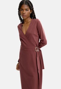 Bordowa sukienka wrap z długimi rękawami, głębokim dekoltem w serek i srebrnym akcentem w talii. Gładka tekstura, o długości do połowy uda.