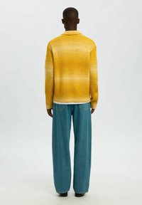 Geltonas megztinis su gradiento raštu, aukštu apykakle ir briaunuota apačia, derinamas su šviesiai mėlynais plačiomis kojomis džinsais ir juodais batais.