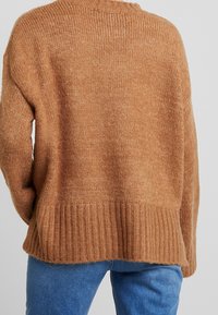 Bruine gebreide trui met een geribbelde zoom, ronde hals en drop shoulder ontwerp, met een zachte textuur en een losse pasvorm. Achteraanzicht weergegeven.