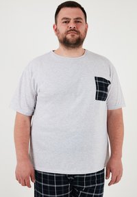 Siva pamučna majica s okruglim izrezom i kratkim rukavima, koja ima crnu kariranu džepu s lijeve strane.
