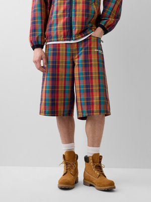 Homme portant une veste à carreaux colorée et un short assorti jusqu'aux genoux, accompagné de chaussettes blanches et de bottes lacées couleur sable.