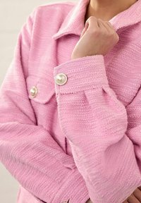 Close-up of a pink textured jacket with pearl buttons on the cuff and pocket, worn by a person with one hand near the collar.