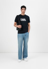 Marineblå bomull T-skjorte med hvit "KARL LAGERFELD" logo, kombinert med lyseblå vide jeans og hvite joggesko, accessorert med en svart veske.