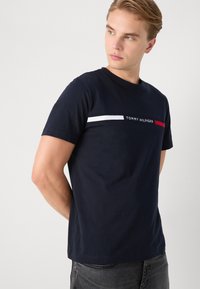 Fiatal férfi sötétkék Tommy Hilfiger pólóban, fehér és piros csíkokkal, balra néz, egyszínű világos háttér előtt.