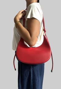 Czerwona skórzana torebka o zaokrąglonym kształcie, z regulowanym paskiem i gładką fakturą. Nosi ją osoba w białej koszuli i ciemnej spódnicy.