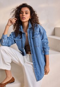 Camicia in denim azzurro chiaro con maniche lunghe, due tasche sul petto e bottoni a pressione. Indossata sopra un top marrone e abbinata a pantaloni bianchi a gamba larga.