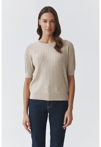 Beige bordás pulóver rövid ujjal, kerek nyakkivágással és texturált anyaggal, sötétkék farmerrel párosítva.