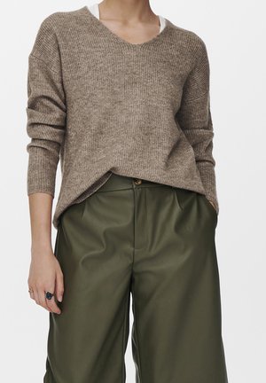 Personne portant un pull marron côtelé à col en V par-dessus un sous-vêtement blanc et un pantalon en cuir vert olive taille haute, les mains dans les poches.