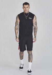 Musta hihaton paita ja mustat shortsit, joissa on punainen vetoketjutarkenne. Malli pukeutunut valkoisiin sukkiin ja mustiin lenkkareihin, seisoo vaalealla taustalla.