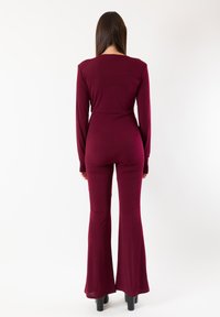 Tuta bordeaux a maniche lunghe con corpetto aderente e gambe svasate, realizzata in morbido tessuto elasticizzato dalla texture liscia.