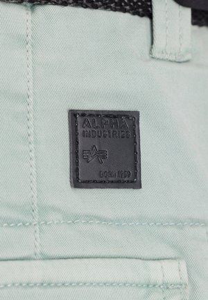 Parche de cuero negro con texto en relieve "ALPHA INDUSTRIES" y "BORN 1959" sobre una tela ligera de color menta. Detalles de costura visibles alrededor del parche.