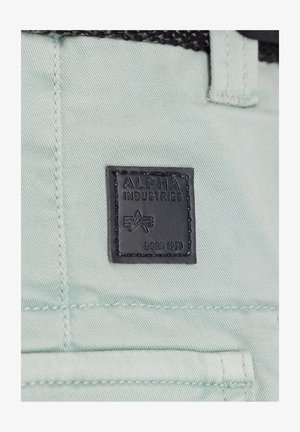 Fascia in pelle nera con testo impresso "ALPHA INDUSTRIES" e "BORN 1959" su tessuto verde menta chiaro. Dettagli di cucitura visibili attorno alla fascia.