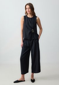 Top senza maniche nero con dettaglio a nodo laterale e rifinitura dorata, abbinato a pantaloni neri larghi a vita alta e scarpe nere basse.