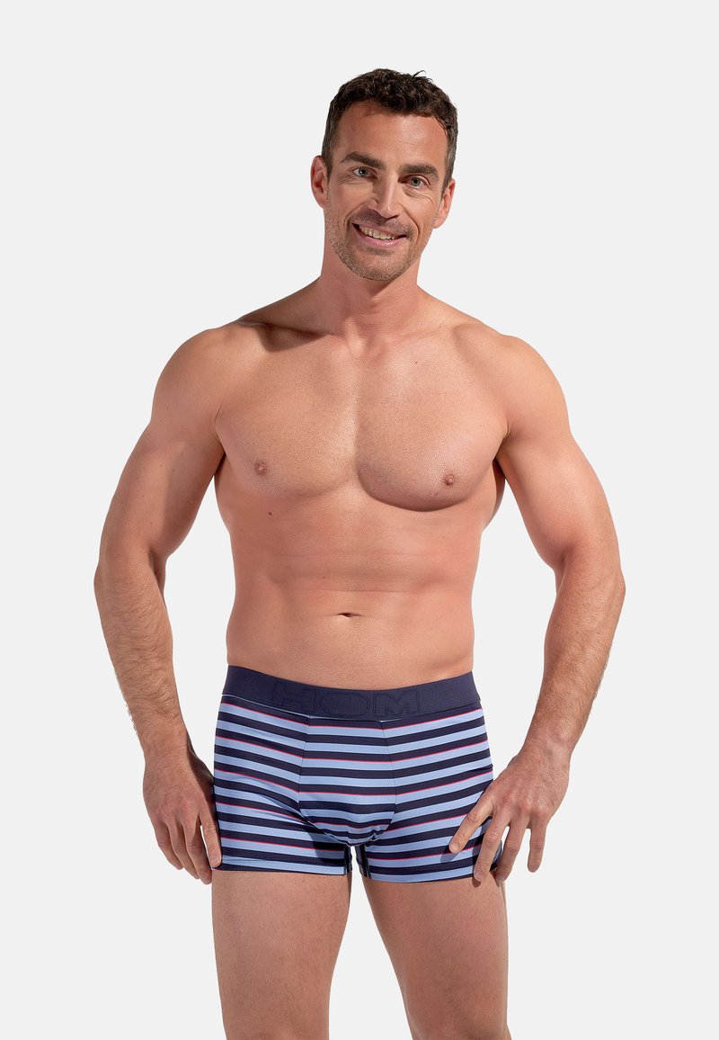 Lachende shirtloze man met kort donker haar, gekleed in blauw en marineblauw gestreepte boxershorts, poserend met handen bij de heupen tegen een effen achtergrond.