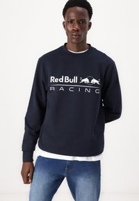 Tamno plavi dukserica s „Red Bull Racing“ logotipom u bijeloj boji, okrugli izrez, duge rukave i materijal od pamuka. Nosena preko bijele majice.