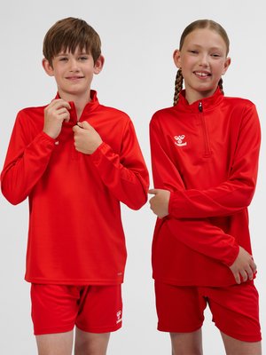 To børn i rødt sportstøj med langærmede lynlåstrøjer og shorts, stående mod en ensfarvet baggrund, smilende.
