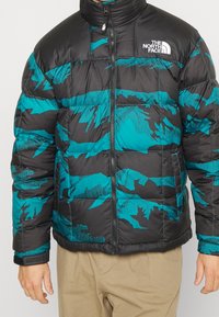 Giacca a vento nera e teal, con motivo mimetico, colletto alto e chiusura con zip. Il materiale sembra liscio e isolante.