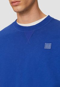 Felpa blu con collo a costine, tessuto strutturato e un piccolo logo azzurro sulla parte sinistra del petto. Design semplice, vestibilità casual.