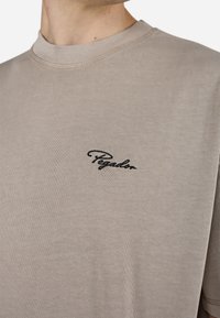 Beige bomull T-skjorte med ribbet rund hals og korte ermer, med svart brodert "Pegador"-logo på brystet. Glatt tekstur.