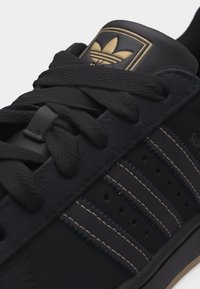 Μαύρο αθλητικό παπούτσι Adidas με σουέντ υφή, μαύρα κορδόνια, τρεις ραμμένες ρίγες στο πλάι και χρυσό λογότυπο Adidas στη γλώσσα.