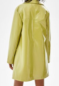 Glanzende limegroene jas met een gestructureerde kraag, lange mouwen en een lengte tot halverwege de dij. Gladde textuur en zichtbare stikseldetails op de naden.