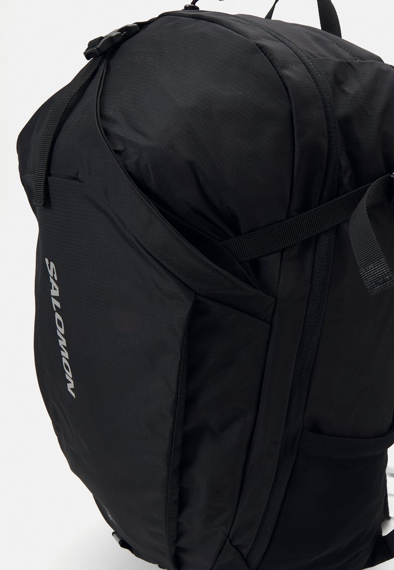 Sac à dos noir Salomon avec un tissu texturé, une forme courbée, une poche latérale et des bretelles ajustables. Présente un logo proéminent d'un côté.