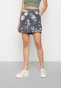 Žena v námořnických květovaných šortkách a olivově zeleném crop topu s bílými platformovými teniskami, stojící proti jednobarevnému bílému pozadí.