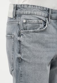 Κοντινό πλάνο ξεθωριασμένου γκρι τζιν με λευκό πουκάμισο, που δείχνει τις μπροστινές και πίσω τσέπες με μεταλλικά τρουκς και θηλιές ζώνης.