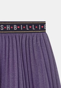 Lila glittrig tyllkjol med svart elastiskt midjeband som har vit och rosa text, samt guldaccentdetaljer.