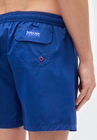 Μπλε μαγιό βερμούδα με πίσω τσέπη που έχει κόκκινο μεταλλικό κρικάκι και ετικέτα με λογότυπο. Το ύφασμα είναι απαλό με ελαστική μέση για άνεση.