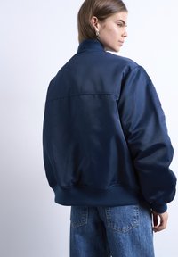 Γυναίκα με κοντά καφέ μαλλιά που φοράει ασημένια κρίκους σκουλαρίκια, μια ναυτικού μπλε μπουφάν τύπου bomber και μπλε τζιν, στραμμένη πλάγια σε ανοιχτό φόντο.