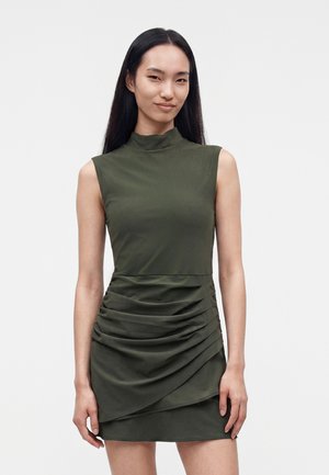 Femme aux longs cheveux lisses portant une robe olive sans manches, col montant, avec une jupe froncée, debout devant un fond uni.