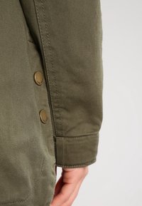 Olivgrön jackärm med synliga gyllene knappar, textil med struktur och prydlig sömnad, som har en sidoficka och en skräddarsydd passform.