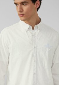 Chemise blanche à manches longues en tissu léger, dotée d'une petite poche et d'une texture subtile. Col traditionnel et fermeture à boutons sur le devant.